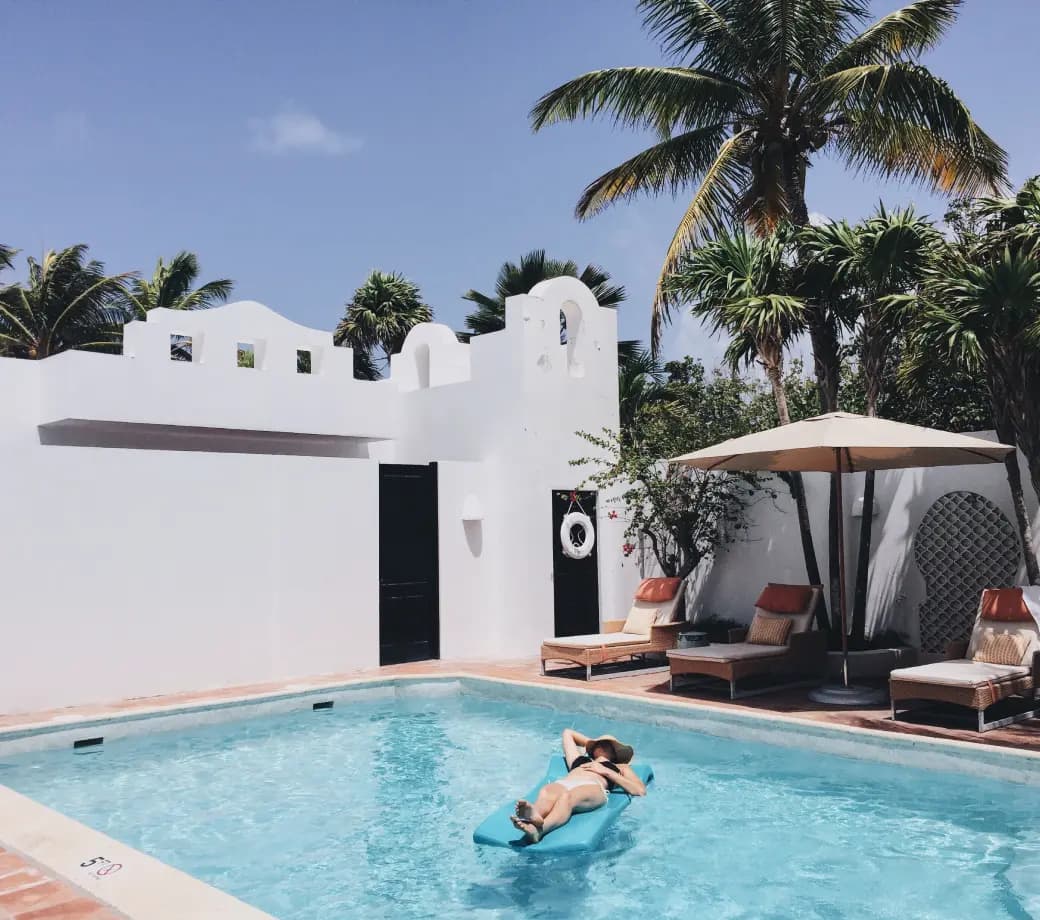 A person relaxes on a floating mat in a turquoise swimming pool, surrounded by white stucco walls and palm trees. Two lounge chairs with umbrellas are nearby, under a clear blue sky.