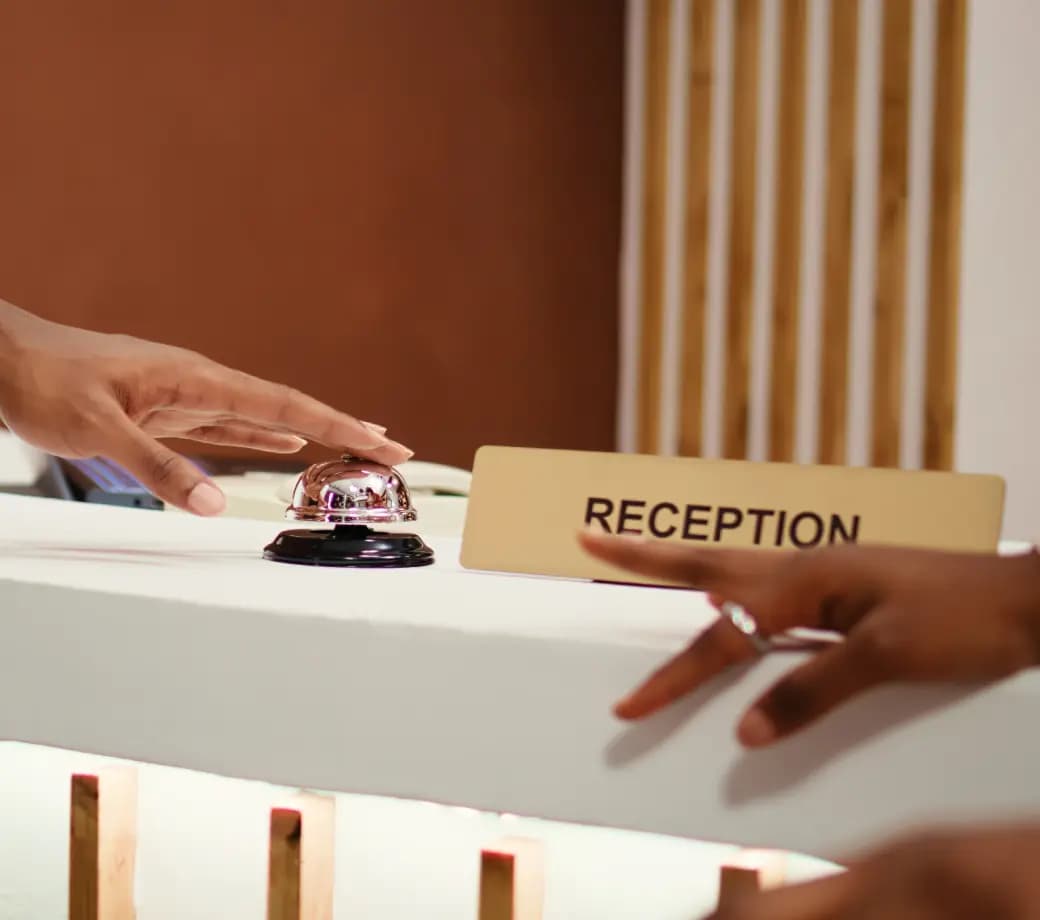 Two hands reaching towards a service bell on a reception desk. A sign labeled Reception is placed next to the bell. The background features a wooden design element.