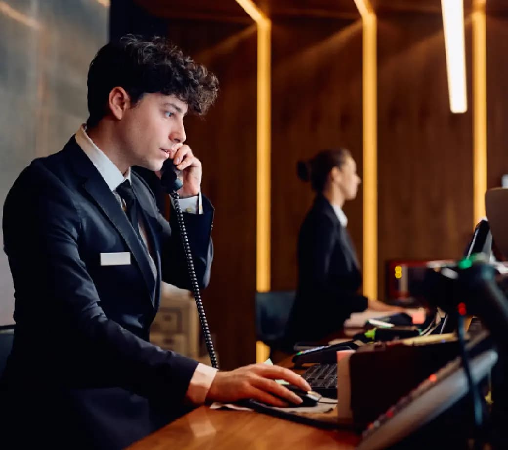 A man in a suit is speaking on the phone while working on a computer at a reception desk. A woman is visible in the background, also working. The room has warm lighting, creating a professional atmosphere.